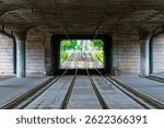 Symmetrical view of empty tram tracks under a concrete bridge leading to green trees