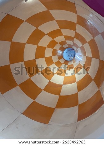 A symmetrical view from the inside of a circular water slide tunnel featuring a bold orange and white checkerboard pattern. The tunnel narrows toward the center, drawing attention to a small safety si