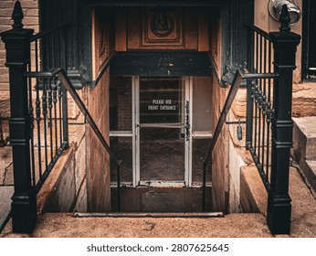 Symmetrical view looking down stone stairs toward a basement glass door with a notice reading please use front entrance between black iron railings