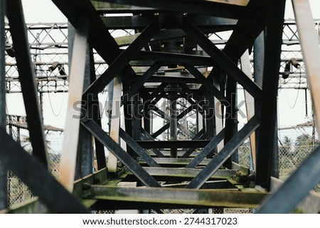 Symmetrical view looking through the geometric steel lattice structure of an electrical power transmission tower. The intersecting metal beams create a striking industrial pattern and leading lines towards the center.