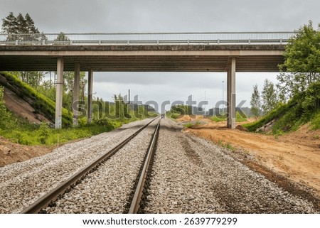 Symmetrical view of railway tracks stretching into the distance and passing beneath a concrete bridge, surrounded by forested terrain and cloudy skies.