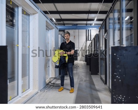 Symmetrical view of a warehouse worker centred in an aisle of stored products, reflective jacket in hand.