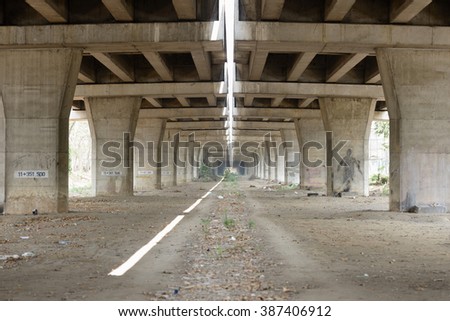 Symmetrical wide-angle view from beneath a massive concrete highway overpass showing a perspective line of thick structural pillars and beams. This industrial architectural landscape highlights the brutalist design of modern infrastructure, with a dirt ground and a thin sliver of natural sunlight filtering through the center gap. The image captures the scale and raw texture of civil engineering, making it ideal for themes related to urban decay, construction, transportation, city planning, and structural engineering. This atmospheric shot under a bridge provides a sense of depth and leading lines, perfect for cinematic backgrounds, urban exploration articles, infrastructure development reports, and gritty city-themed marketing materials. The neutral grey tones and repetitive geometric patterns offer a strong visual foundation for architectural presentations and editorial content about metropolitan expansion.