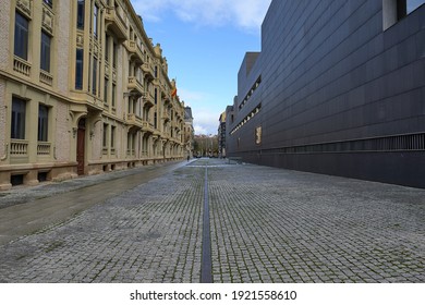 Symmetry And Contrast Between Two Buildings, One With A Smooth Black Wall, The Other With A Light Brick Wall, And People Walking On The Cobbled Street