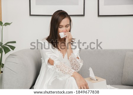 Symptoms of allergic rhinitis in women. Sick woman in white nightwear with a cold blowing her nose into a tissue paper at home. Cold weather allergies