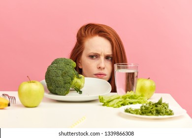 Symptoms Of Anorexia Manifested In Aversion To Food. Portrait Of Grimacing Unsatisfied Facial Emotional Expression Lady Refusing To Eat Fresh Green Salad In Bowl On Table In Light Room