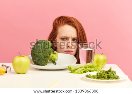 Symptoms of anorexia manifested in aversion to food. Portrait of grimacing unsatisfied facial emotional expression lady refusing to eat fresh green salad in bowl on table in light room