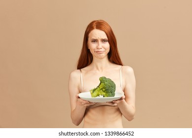 Symptoms of anorexia manifested in disgust for food. Portrait of grimacing unsatisfied facial emotional expression young woman refusing to eat fresh broccoli on plate, posing over pink background