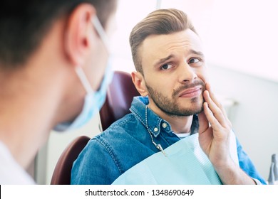 Symptoms Of Gums Pain. A Man With A Worried Face Is Holding His Hand On His Cheek  Because Of Irritating Pain In Front Of A Dentist Who Is Going To Give A Patient A Treatment.