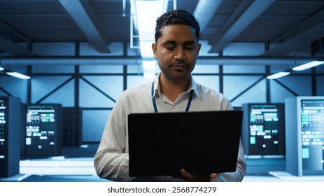 System administrator in data center using notebook to design, implement and manage gear, ensuring optimal performance and reliability. Indian man in server room coding on laptop, camera B