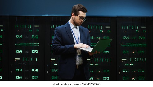 System administrator in glasses and suit standing in the middle of server room holding laptop and enters data into database inspecting technical characteristics of device. - Powered by Shutterstock