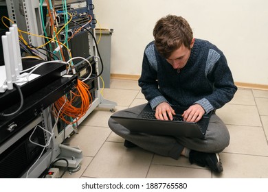 A System Administrator With A Laptop Works In A Data Center. A Teenager Programs At The Server Racks. Young Guy Is Sitting On The Floor In The Server Room.