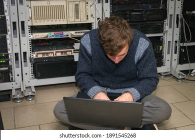 A System Administrator With A Laptop Works In A Data Center. A Teenager Programs At The Server Racks.Young Guy Is Sitting On The Floor In The Server Room.