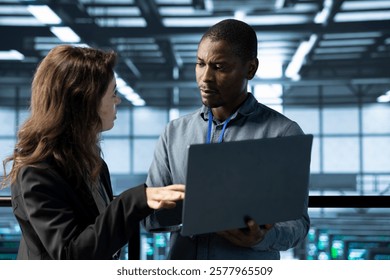 System administrators in data center using laptop, analyzing metrics, optimizing storage volumes. Server farm IT experts using notebook, talking, ensuring seamless operations