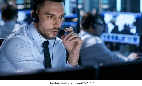 In System Control Center Technical Support Specialist Speaks into Headset. His Colleagues are Working in the Background in a Room Full of Screens.