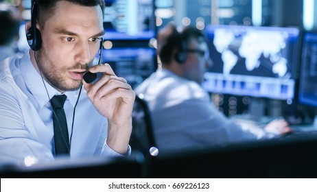 In System Control Center Technical Support Specialist Speaks Into Headset. His Colleagues Are Working In The Background In A Room Full Of Screens.