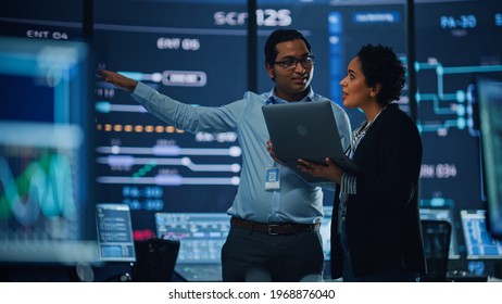 In the System Control Room Project Manager and IT Technical Engineer with Laptop Have Discussion, surrounded by Multiple Monitors with Graphics. Big Monitor Shows Interactive Server Blockchain Info.