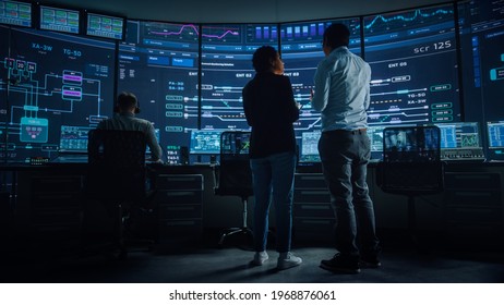 In The System Control Room Project Manager And IT Technical Engineer Have Discussion, They're Surrounded By Multiple Monitors With Graphics. Big Monitor Shows Interactive Server Blockchain Info.