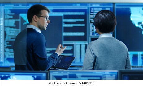 In the System Control Room IT Specialist and Project Engineer Have Discussion while Holding Laptop, they're surrounded by Multiple Monitors with Graphics. They Work in a Data Center on Data Mining.
