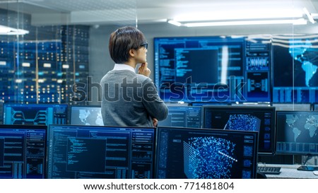 In the System Control Room Technical Operator Stands and Monitors Various Activities Showing on Multiple Displays with Graphics. Administrator Monitors Work of  Artificial Intelligence.
