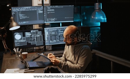 IT system engineer comparing data files on clipboard papers and multiple monitors to program new server interface. Developing html script on terminal window, coding database. Handheld shot.