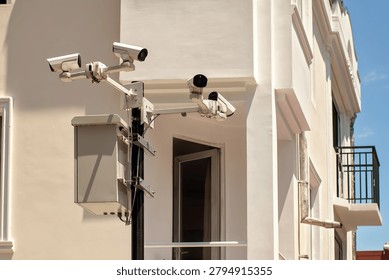 System of four white CCTV security cameras and a control box mounted on a metal pole on the background of a modern residential building facade.