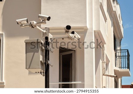 System of four white CCTV security cameras and a control box mounted on a metal pole on the background of a modern residential building facade.