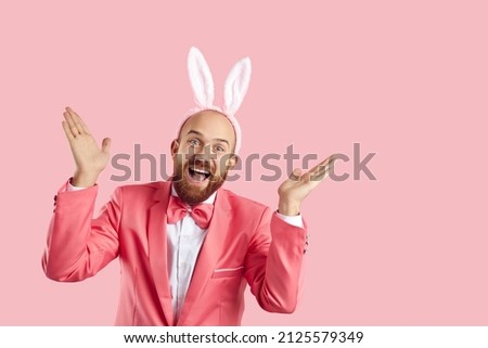 Ta da it's me aren't you surprised. Happy guy in bunny costume having fun on Easter Day. Excited smiley goofy bearded man wearing suit, bowtie and funny ears playing peekaboo on pink studio background