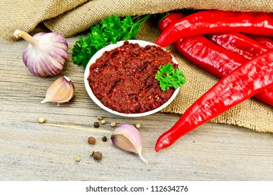 Tabasco in a white cup, fresh red peppers, garlic, peppercorns, mustard seeds on the background burlap and wooden planks