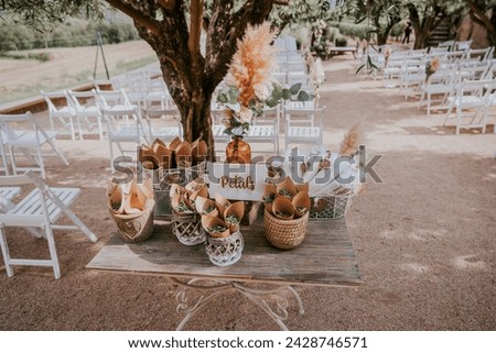 TABLE WITH BASKETS WITH RICE AND PETALS FROM A WEDDING. THERE IS A SIGN THAT SAYS PETALS. IN THE BACKGROUND ARE THE WHITE CHAIRS OF A WEDDING CEREMONY. THE PETALS ARE GREEN OLIVE LEAVES.
