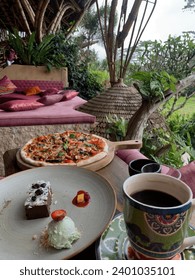 Table in cafe with pizza, chocolate dessert brownie with ice cream, and a large cup of coffee. In the background are a pink sofa with pillows, a thatched roof, tree trunks and green grass.