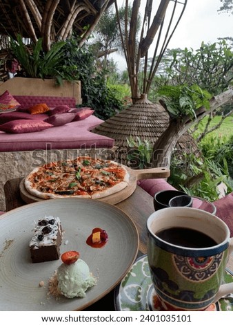 Table in cafe with pizza, chocolate dessert brownie with ice cream, and a large cup of coffee. In the background are a pink sofa with pillows, a thatched roof, tree trunks and green grass.
