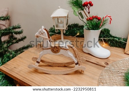 Table with Christmas decor. Decorative light pole in the background. Ceramic boot with floral arrangements and candy cane inside. Wooden seesaw horse at the front.