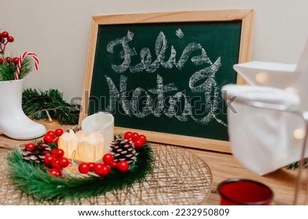 Table with Christmas decor. On the green board merry christmas is written in portuguese. Ceramic boot with floral arrangements and candy cane inside. Garland with candles in the middle.