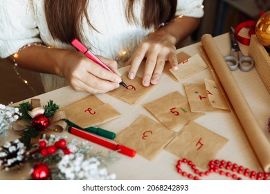 A Table With Christmas Decorations And The Hands Of A Girl Writing Numbers On An Advent Calendar Diy. High Quality Photo