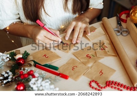 a table with Christmas decorations and the hands of a girl writing numbers on an advent calendar diy. High quality photo