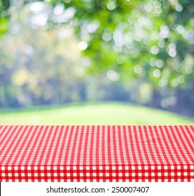 Table With Cloth For Picnic Background, Empty Red And White Tablecloth Over Blur Green Tree Park Garden And Abstract Bokeh In Spring, Summer Background, For Food, Product Display Montage Template