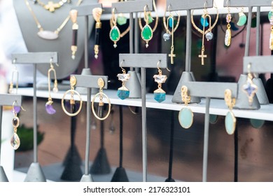 A Table Of Decorative And Colorful Cosmetic Earrings.  The Glamourous Balls,  Jewelry, And Fashionable Earrings Are Hanging On Display At A Bazaar. The Gold Trinkets Have Multiple Colored Stones.