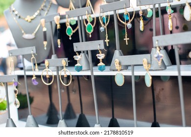 A Table Of Decorative And Colorful Cosmetic Earrings.  The Glamourous Balls,  Jewelry, And Fashionable Earrings Are Hanging On Display At A Bazaar. The Gold Trinkets Have Multiple Colored Stones.