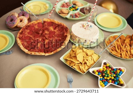Table displaying pizza, cake, donuts, cookies, chips, fries, candy and empty plates arranged for party celebration, food items placed on pastel colored dishes ready for serving