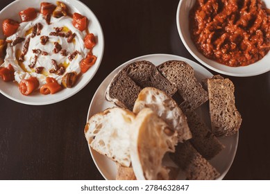 A table displays various types of bread alongside bowls of tomato dip and yogurt with herbs. This food arrangement shows a simple meal shared among friends.