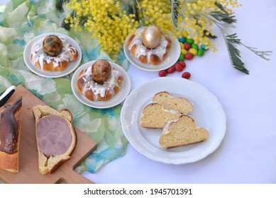 Table With Easter Food: Sliced Easter Ham In Sweet Dough On Wooden Board, Slices Of Braided Bread With Sugar Glaze And Easter Cakes In A Shape Of Easter Nest With An Egg In Background 