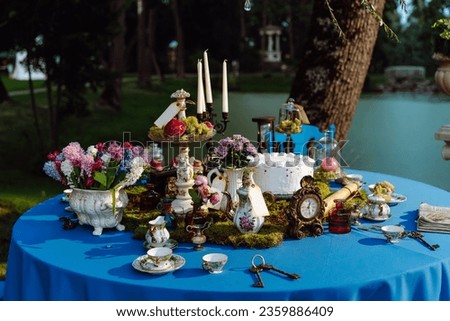 Table is elegantly decorated for tea party in style of Alice in Wonderland. Cups and saucers, candles in candlestick, cake, old keys, flowers in vase, clock on blue tablecloth late at night near pond