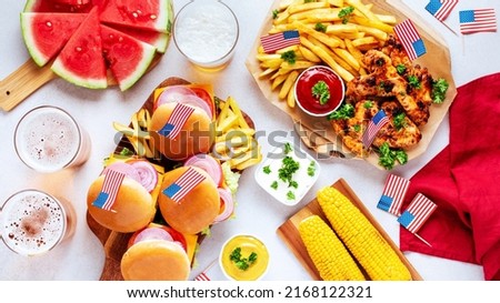 Table with food for USA 4th July Independence Day. Fourth July party or picnic food with burgers, chicken wings, corn, fries and beer, decorated with american flags, top view, copy space
