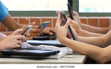 A Table Of High School Students All Holding Multiple Digital Devices Such As Phones And Tablets During A Class Lesson. Contemporary Education And Connected Technology. 