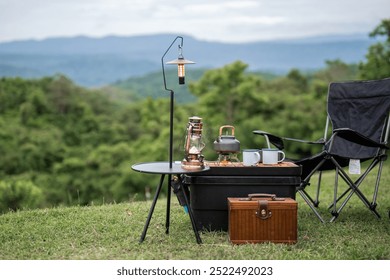 A table with a lantern and a tea kettle on it, and a black chair next to it. The scene is set in a grassy field with mountains in the background. The table and chair are set up for a camping trip