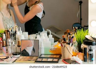 A Table With Make-up Artist Tools With A Large Hollywood Mirror With Light Bulbs Reflecting A Blurred Image Of A White Makeup Artist Applying Makeup To A White-skinned Model With Blond Hair