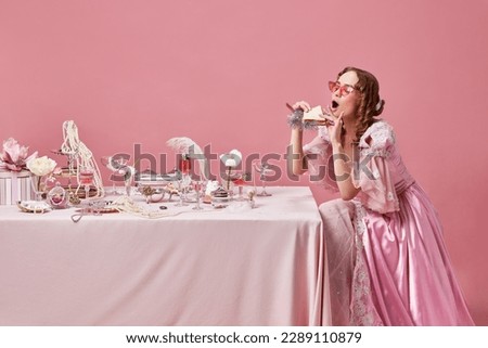 Table manners. Portrait of beautiful, hungry, blond princess wearing fancy pink dress and sunglasses eating sandwich over studio background. Concept of medieval, beauty, healthy food, baroque, ad