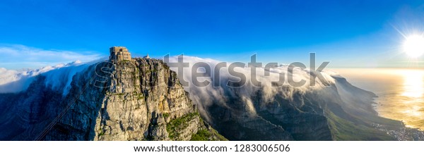 Table Mountain cable\
way covered in clouds sweeping over the twelve apostles during\
sunset in Cape Town