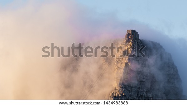 Table Mountain cable\
way covered in clouds sweeping over the twelve apostles during\
sunrise in Cape Town